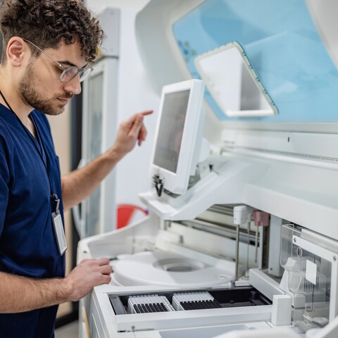 Ein Laborfachmann bedient ein automatisiertes Analysegerät und überprüft Proben in einem Labor. Das Gerät verfügt über ein integriertes Display und Probenhalter.