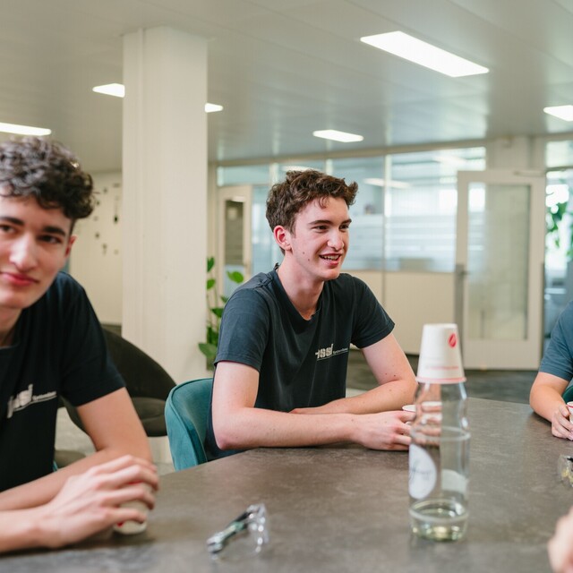 Jossi apprentices sit together around a table, chatting in a relaxed atmosphere. The scene highlights teamwork, young talent development, and an open workplace culture.