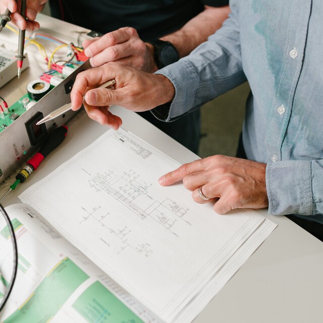 Zwei Personen prüfen einen Schaltplan und arbeiten gemeinsam an einer geöffneten elektronischen Steuerung. Messgeräte, Werkzeuge und Leiterplatten deuten auf Fehlersuche, Inbetriebnahme oder Wartung einer industriellen Anlage hin.