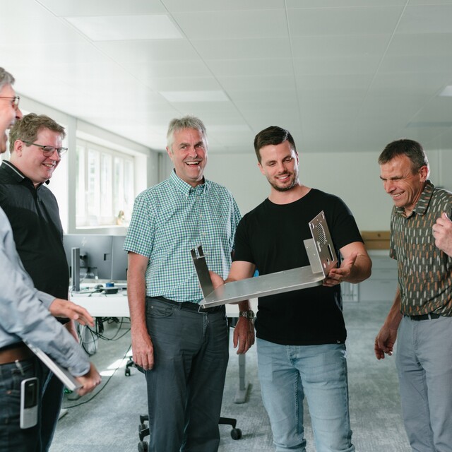 Eine Gruppe von Jossi-Mitarbeitenden begutachtet gemeinsam ein mechanisches Bauteil und diskutiert dessen Konstruktion. Die Szene vermittelt Teamarbeit, Wissensaustausch und Zusammenarbeit in einer technischen Entwicklungs- oder Produktionsumgebung.
