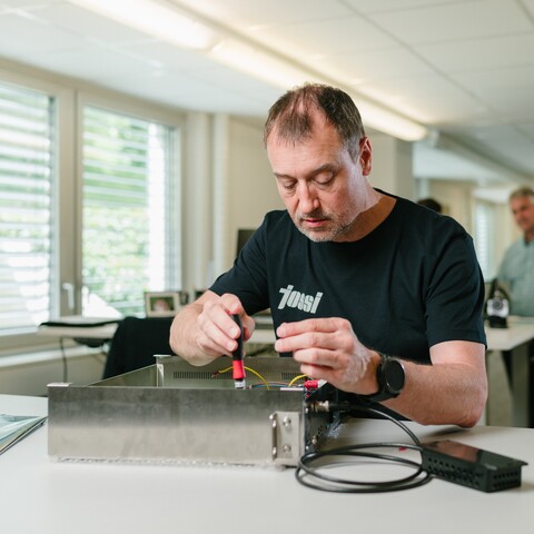 A Jossi employee assembles and wires electronic components of a prototype using a screwdriver. The scene illustrates the precise assembly and testing of an industrial automation solution.