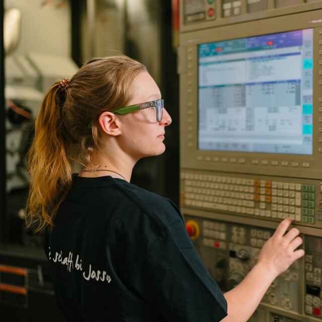 Eine Jossi-Mitarbeiterin bedient das Steuerungspanel einer Fertigungsmaschine und überwacht den Produktionsprozess. Die Szene zeigt die präzise Steuerung und Kontrolle einer industriellen Automatisierungsanlage.