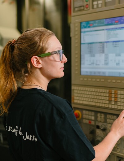 Eine Jossi-Mitarbeiterin bedient das Steuerungspanel einer Fertigungsmaschine und überwacht den Produktionsprozess. Die Szene zeigt die präzise Steuerung und Kontrolle einer industriellen Automatisierungsanlage.