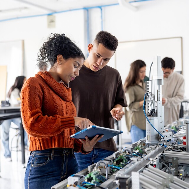 Zwei Studierende arbeiten gemeinsam an einer automatisierten Versuchsanlage und betrachten die Steuerung auf einem Tablet. Weitere Personen arbeiten im Hintergrund des Labors.