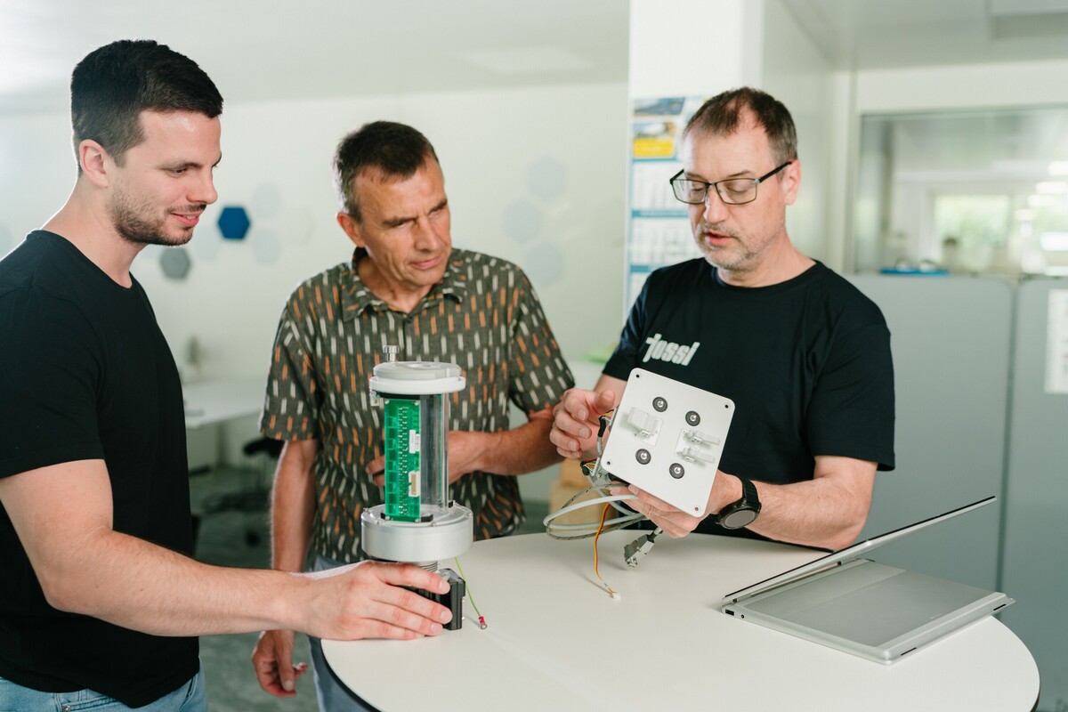 Drei Jossi-Mitarbeiter besprechen den Aufbau eines technischen Prototyps und prüfen elektronische Komponenten. Die Szene zeigt den gemeinsamen Austausch und die Entwicklung einer industriellen Automatisierungslösung.