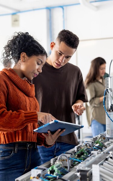 Zwei Studierende arbeiten gemeinsam an einer automatisierten Versuchsanlage und betrachten die Steuerung auf einem Tablet. Weitere Personen arbeiten im Hintergrund des Labors.