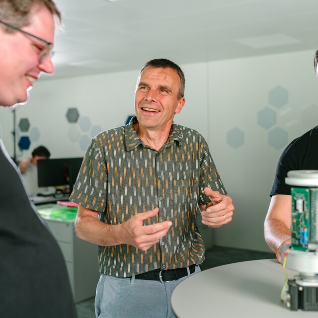 Drei Jossi-Mitarbeiter tauschen sich vor einem technischen Prototypen über dessen Entwicklung und Funktion aus. Die Szene vermittelt Zusammenarbeit, Erfahrungsaustausch und gemeinsame Produktentwicklung im Unternehmen.