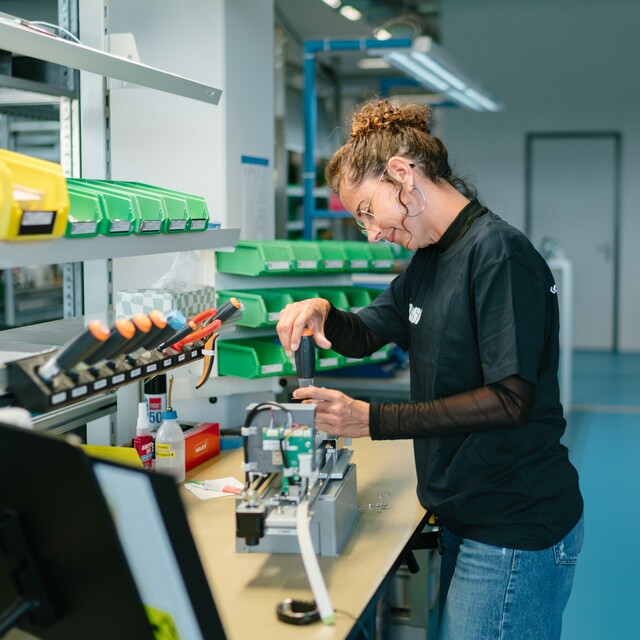 Eine Jossi-Mitarbeiterin montiert und prüft eine Baugruppe an einem modernen Montagearbeitsplatz. Die Szene zeigt präzise Fertigungsarbeit und Qualitätsbewusstsein bei der Herstellung industrieller Automatisierungslösungen.