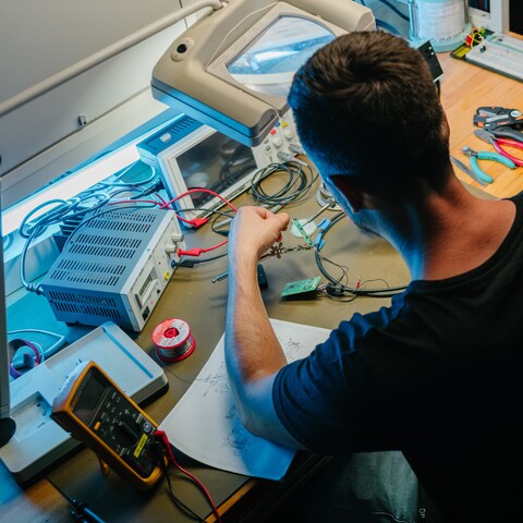 A Jossi employee tests and inspects an electronic assembly at an electronics workstation equipped with measuring instruments and soldering tools. The scene highlights development, testing, and validation work for industrial automation solutions.