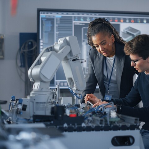 An instructor supports a student while working on a robotic cell in a modern automation laboratory. The scene highlights hands-on training, knowledge transfer, and industrial robotics.