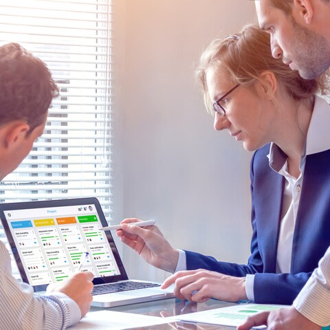 Three professionals review project progress using a digital Kanban board displayed on a laptop. The scene represents agile project planning, collaboration, and digital task management.