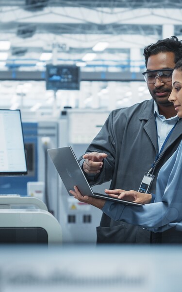 Zwei Ingenieur:innen besprechen Produktionsdaten auf einem Laptop in einer modernen Halbleiterfertigung. Im Hintergrund sind Fertigungsanlagen und ein Monitor mit Analyseinformationen zu sehen.