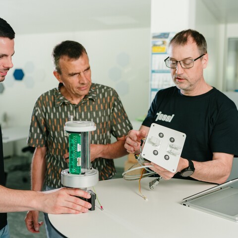 Three Jossi employees discuss the design of a technical prototype while inspecting electronic components. The scene highlights collaboration and the development of an industrial automation solution.