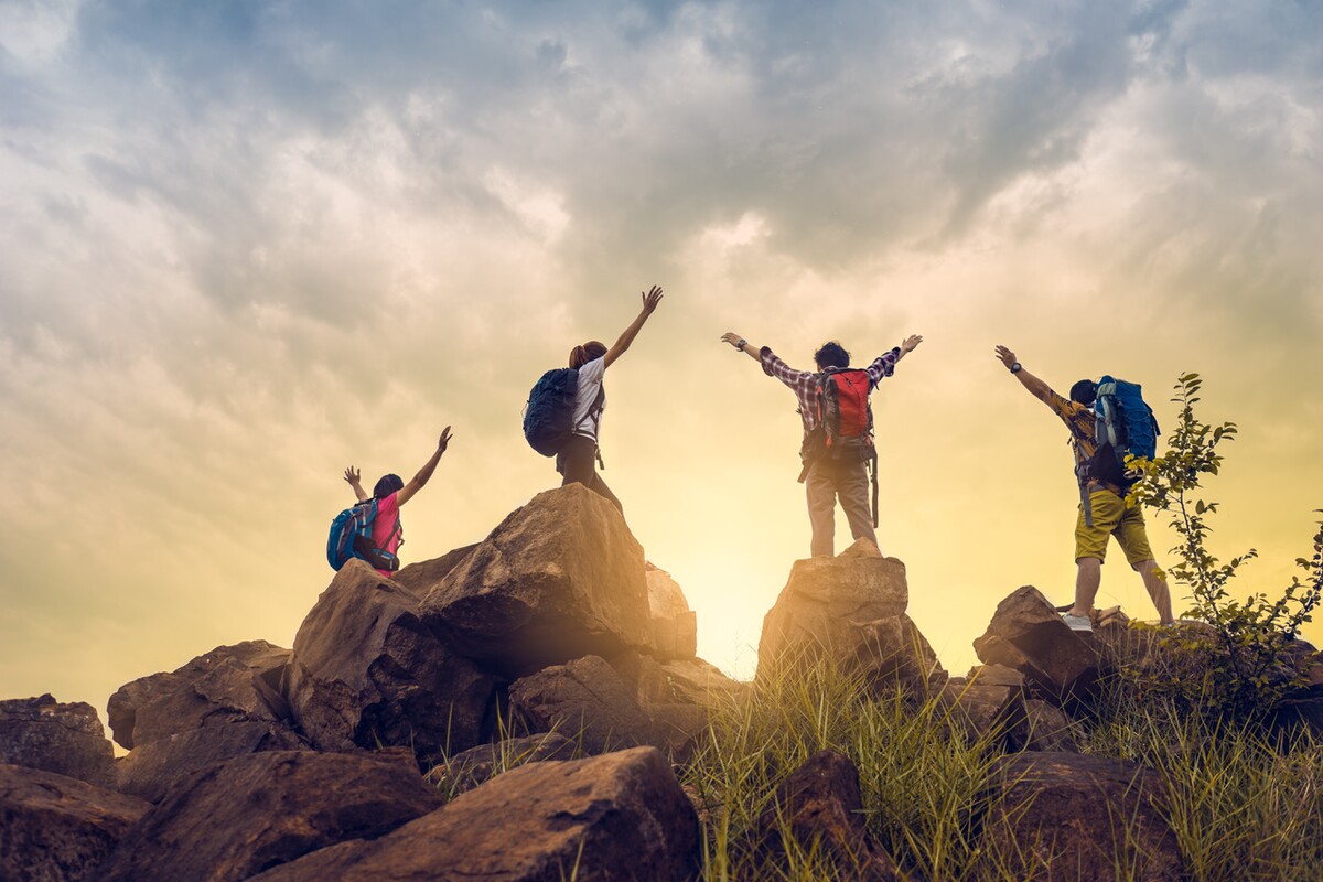 Four people stand on rocky outcrops with their arms outstretched, facing the sunrise. The scene symbolizes teamwork, unity, and achieving goals together.