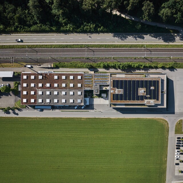 Top-down aerial view of the Jossi company site featuring production and office buildings, rooftop solar panels, and the surrounding transport links and green spaces. The image provides an overview of the company campus and its modern infrastructure.