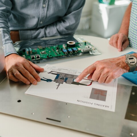 Two professionals review the design of an electronic assembly using a technical drawing. A circuit board, metal enclosure, and additional components for assembly or commissioning are laid out on the table.