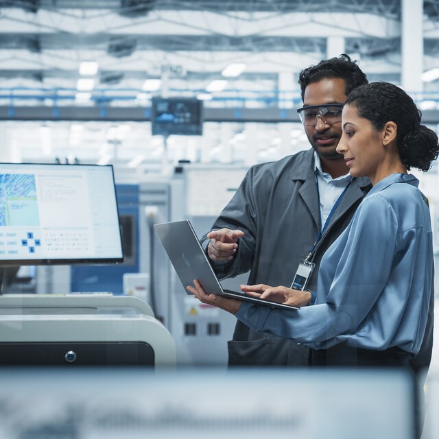 Zwei Ingenieur:innen besprechen Produktionsdaten auf einem Laptop in einer modernen Halbleiterfertigung. Im Hintergrund sind Fertigungsanlagen und ein Monitor mit Analyseinformationen zu sehen.
