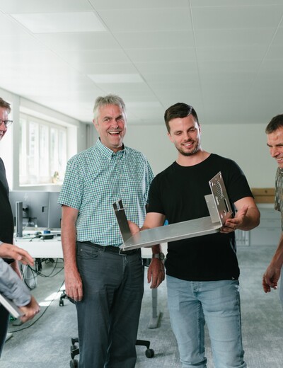 Eine Gruppe von Jossi-Mitarbeitenden begutachtet gemeinsam ein mechanisches Bauteil und diskutiert dessen Konstruktion. Die Szene vermittelt Teamarbeit, Wissensaustausch und Zusammenarbeit in einer technischen Entwicklungs- oder Produktionsumgebung.