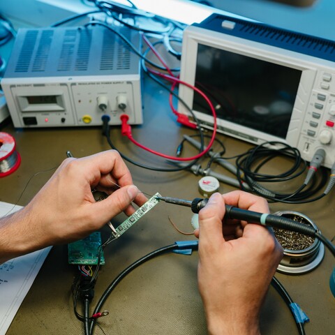 A Jossi employee solders a circuit board at an electronics workstation equipped with test and measurement instruments. The scene highlights precise development and manufacturing work on an industrial automation solution.